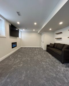 Modern basement living room with gray carpet, dark sofa, wall-mounted TV, electric fireplace, and floating shelves. - GTA